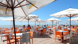 Zona exterior de cafetería con sillas naranjas y blancas, sombrillas y vista al mar en Camping Pra'delle Torri.