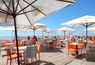 Zona exterior de cafetería con sillas naranjas y blancas, sombrillas y vista al mar en Camping Pra'delle Torri.