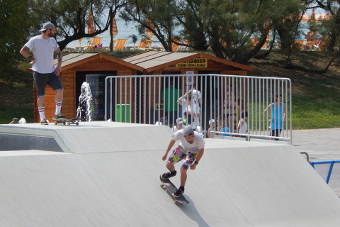 Two skateboarders on a ramp at Camping Pra'delle Torri, with people and a cabin in the background, Veneto, Italy.