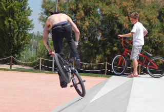 Two people riding BMX bikes in a skatepark at Camping Pra'delle Torri in Veneto, Italy, on a sunny day.