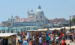 Multitud paseando entre puestos de mercado junto al canal, con edificios históricos y cúpulas al fondo en Venecia.