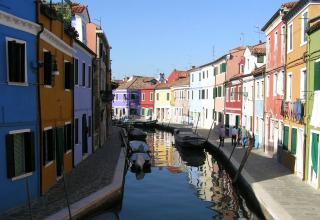 Colorful houses along a canal with boats in Burano, Veneto, Italy, under a clear blue sky.
