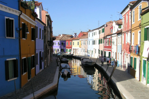 Casas coloridas junto a un canal con barcos en Burano, Véneto, Italia, bajo un cielo azul claro.