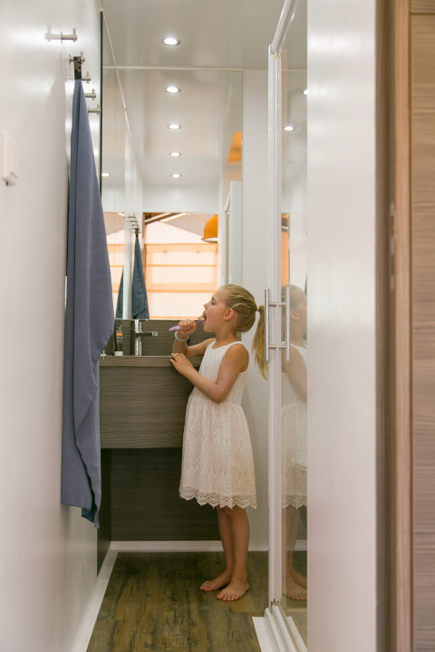 Niña con vestido blanco cepillándose los dientes en un baño estrecho de SunLodge Safari, Camping Pra'delle Torri, Italia.