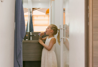 Niña con vestido blanco cepillándose los dientes en un baño estrecho de SunLodge Safari, Camping Pra'delle Torri, Italia.