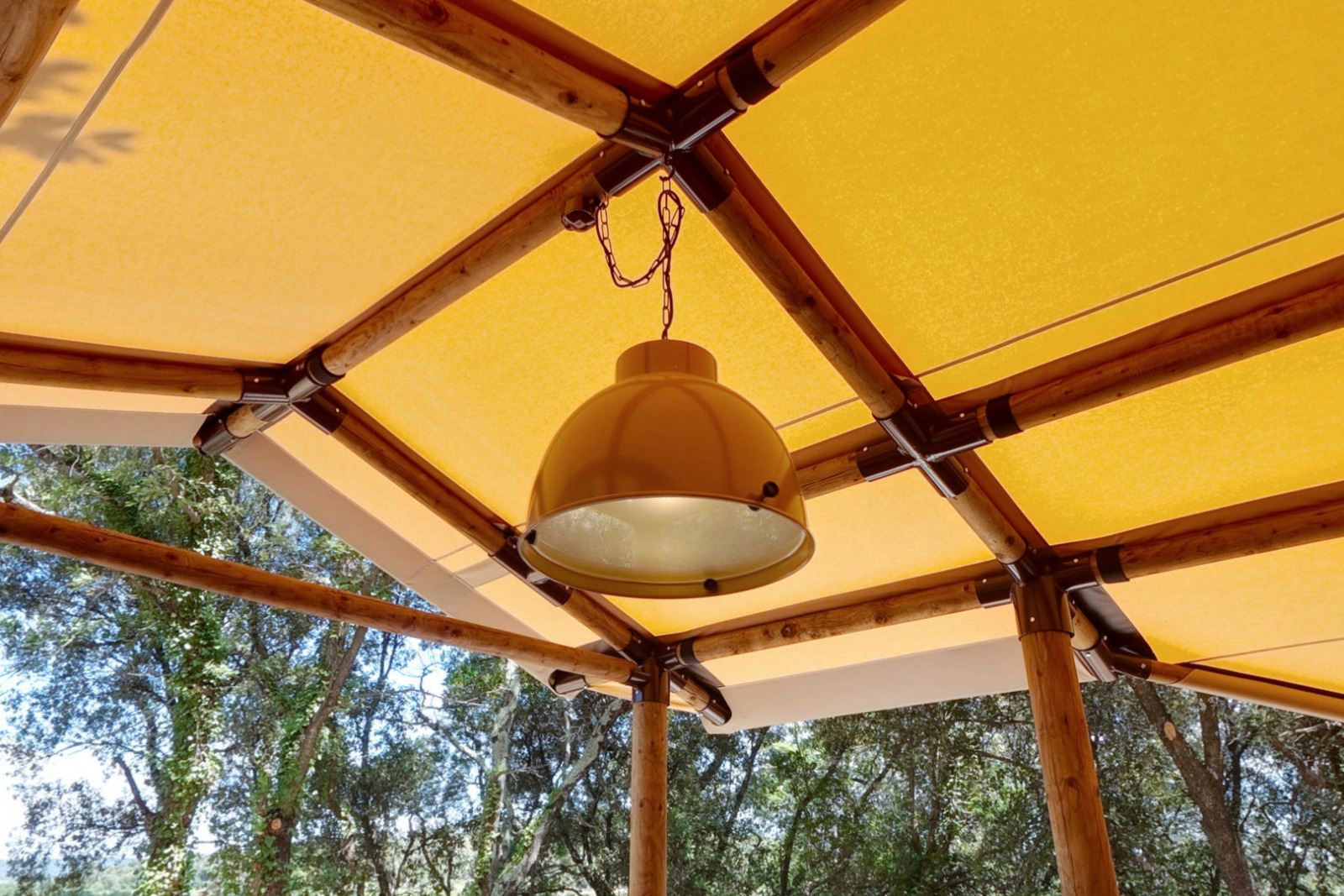 Lamp hangt aan het plafond van een glamping tent met bamboe frame en geel doek, bomen rondom buiten.