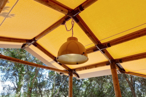 Lamp hangt aan het plafond van een glampingtent met bamboeframe en geel dak, omgeven door natuur.