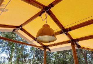 Lampe suspendue au plafond d’une tente glamping avec armature en bambou et toit jaune, nature autour.