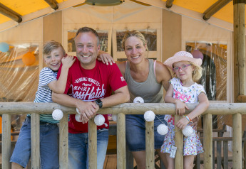 A happy family stands on the veranda of SunLodge Safari at Camping Pra'delle Torri in Italy.