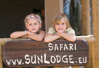 Due bambini appoggiati su un cartello di legno a SunLodge Safari, Camping Pra'delle Torri, Italia.