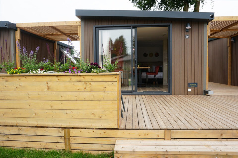 Foto exterior de una cabaña glamping moderna con terraza de madera, macetero y grandes puertas de cristal.