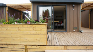 Foto esterna di una moderna cabina glamping con terrazza in legno, fioriera e ampie porte di vetro.