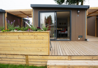 Buitenfoto van een moderne glampinghut met houten terras, bloemenbak en grote schuifdeuren.