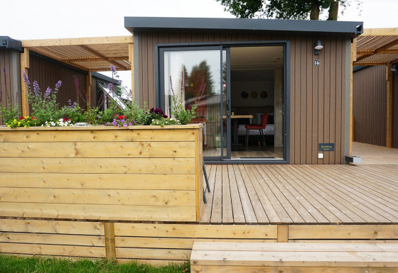 Foto esterna di una moderna cabina glamping con terrazza in legno, fioriera e ampie porte di vetro.