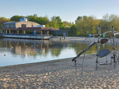 Vista al lago con playa de arena, área de juegos y edificio al fondo en Camping De Gouwe Stek, Holanda.