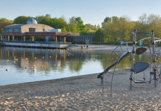 Uitzicht op meer met zandstrand, speeltuin en gebouw op de achtergrond bij Camping De Gouwe Stek, Holland.