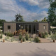 Moderne lodge med træterrasse, havemøbler og omgivet af sand og grønne planter under blå himmel.