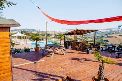 Terraza de madera con mesas, bar y piscina, rodeada de colinas en Agricola La Casetta, Marche, Italia.