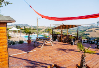 Terraza de madera con mesas, bar y piscina, rodeada de colinas en Agricola La Casetta, Marche, Italia.