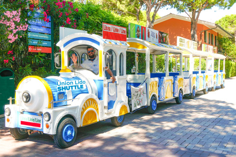 A colorful Union Lido Shuttle train with a smiling driver travels through Camping Union Lido Mare holiday park in Veneto.