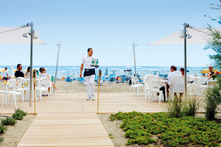 A waiter serves drinks to people relaxing on white plastic chairs by the beach at Union Lido, Veneto, Italy.
