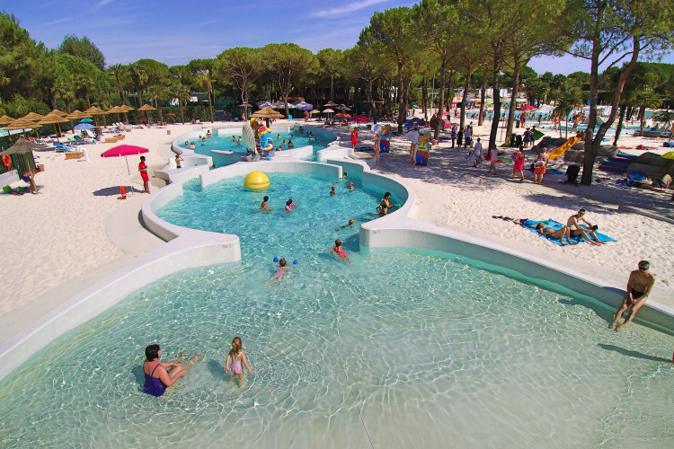 Families enjoy the large pool and sandy beach surrounded by trees at Camping Union Lido Mare in Veneto, Italy.
