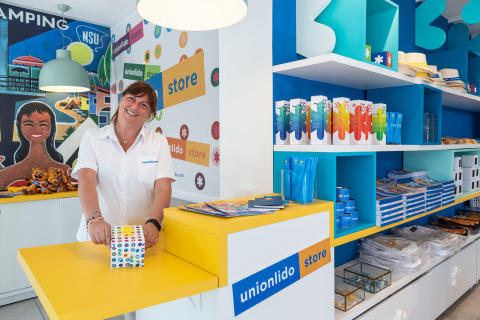 Empleada en la tienda Union Lido de un camping en Véneto, Italia, rodeada de recuerdos y productos coloridos.