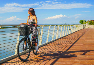 Donna in bicicletta sulla passerella in legno al Camping Union Lido Mare, Veneto, Italia, cielo sereno e sole.