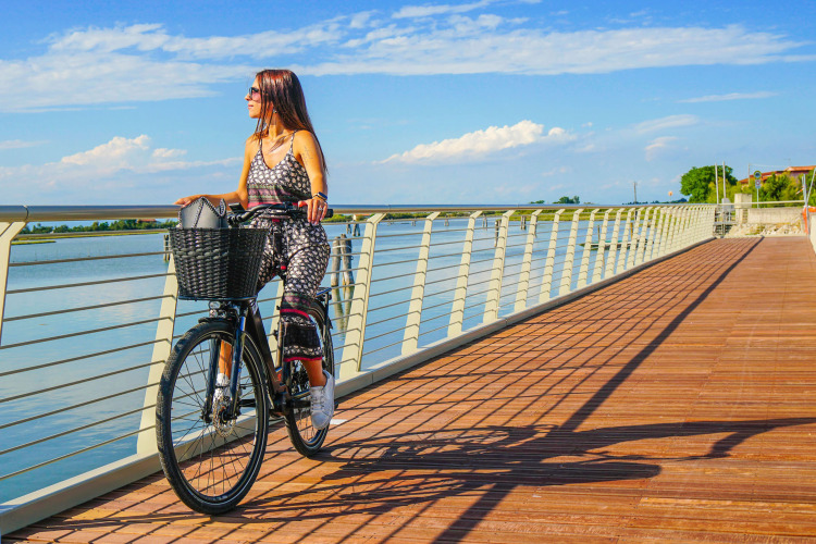 Vrouw fietst op een houten promenade langs het water bij Camping Union Lido Mare, Veneto, Italië, blauwe lucht.