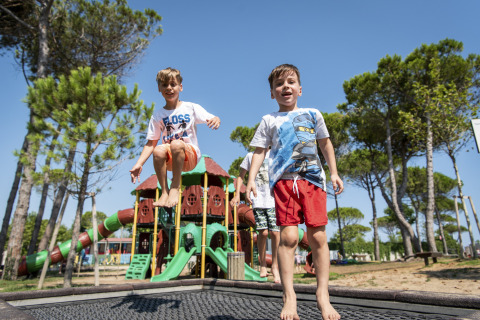 Dos niños juegan y saltan en una cama elástica en el Camping Union Lido Mare en Veneto, Italia, rodeados de árboles.