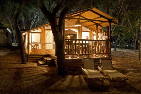 Gezellige, verlichte lodge in het bos 's nachts met personen op het terras en twee ligstoelen vooraan.
