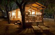 Gezellige, verlichte lodge in het bos 's nachts met personen op het terras en twee ligstoelen vooraan.