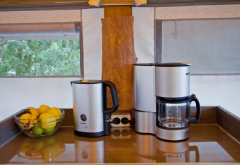 Rincón de cocina glamping con hervidor eléctrico, cafetera y un bol de frutas junto a amplias ventanas.