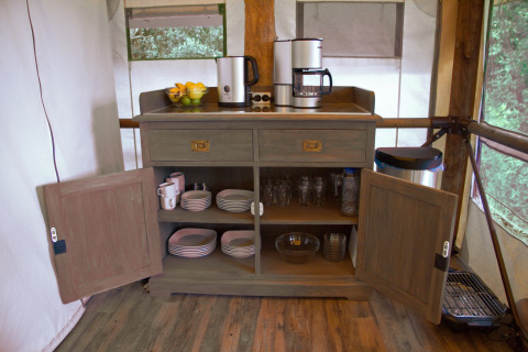 Glamping-keukenhoek met open kast, koffiezetapparaten, borden, glazen, fruit en een afvalbak.