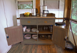 Coin cuisine de glamping avec placard ouvert, cafetières, vaisselle, verres, fruits et poubelle.