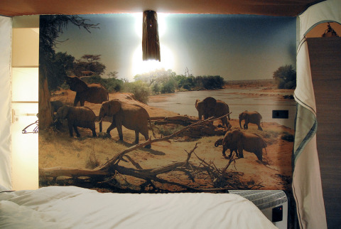 Alojamiento glamping con un gran mural de elefantes junto a un río, cama en primer plano.