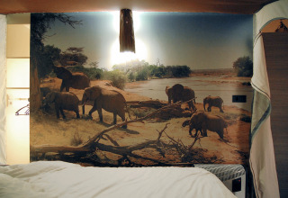 Hébergement glamping avec une grande fresque murale d’éléphants près d’une rivière, lit au premier plan.