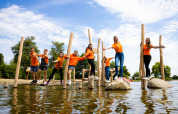 Gruppe unge i orange trøjer leger ved vandet i feriepark med glamping, solrig sommerdag.