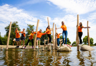 Groep jongeren in oranje shirts speelt op houten klimtoestel in het water bij een glamping vakantiepark.
