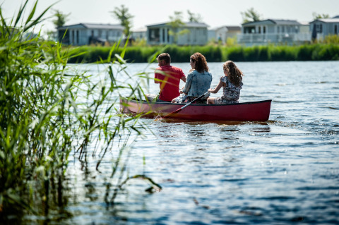 Family paddling a canoe on a lake at a holiday park with glamping cabins and reeds in the background.