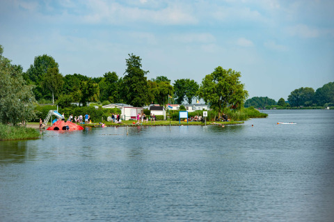 Parco vacanze con glamping sul lago, persone che nuotano e bambini che giocano tra alberi e natura.