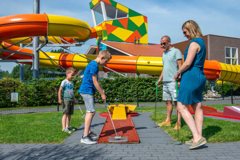 Familia jugando al minigolf frente a toboganes acuáticos coloridos en un parque vacacional con glamping.