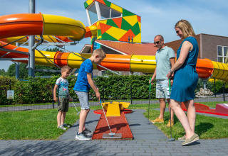 Familia jugando al minigolf frente a toboganes acuáticos coloridos en un parque vacacional con glamping.