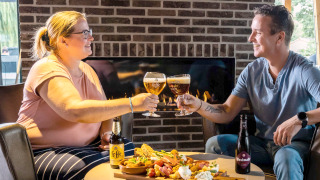 Dos personas brindan con cerveza y disfrutan de aperitivos frente a la chimenea en un parque de glamping.