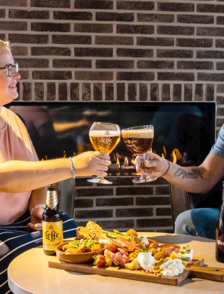 Zwei Personen stoßen mit Bier an und genießen Snacks vor einem Kamin in einem Glamping-Ferienpark.