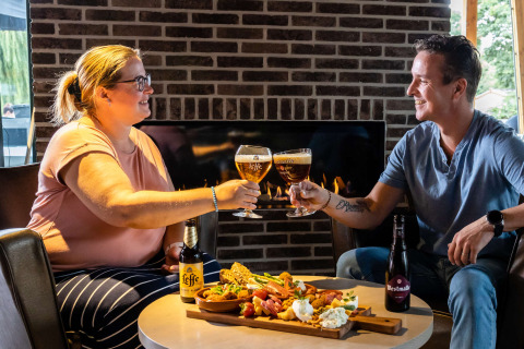 Dos personas brindan con cerveza y disfrutan de aperitivos frente a la chimenea en un parque de glamping.