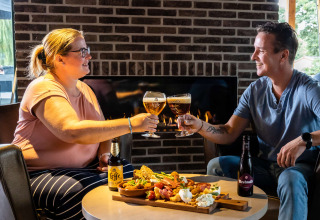 Two people toast with beers and enjoy snacks by a fireplace at a glamping holiday park accommodation.