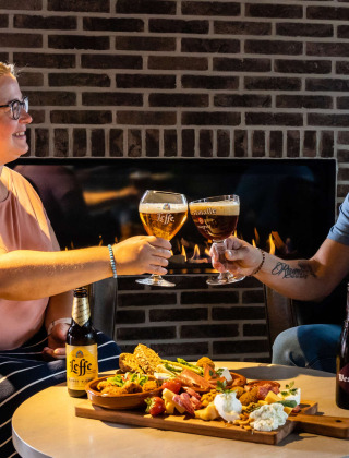 Dos personas brindan con cerveza y disfrutan de aperitivos frente a la chimenea en un parque de glamping.