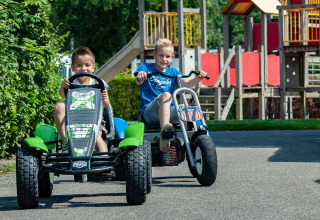 Deux enfants conduisent des véhicules à pédales devant une aire de jeux dans un parc de vacances en glamping.