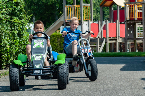 To børn kører på pedaldrevne køretøjer foran en legeplads i en feriepark med glampingmuligheder.